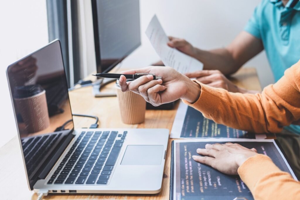 Two software developers at a desk, one pointing a pen at a laptop while the other holds printed code