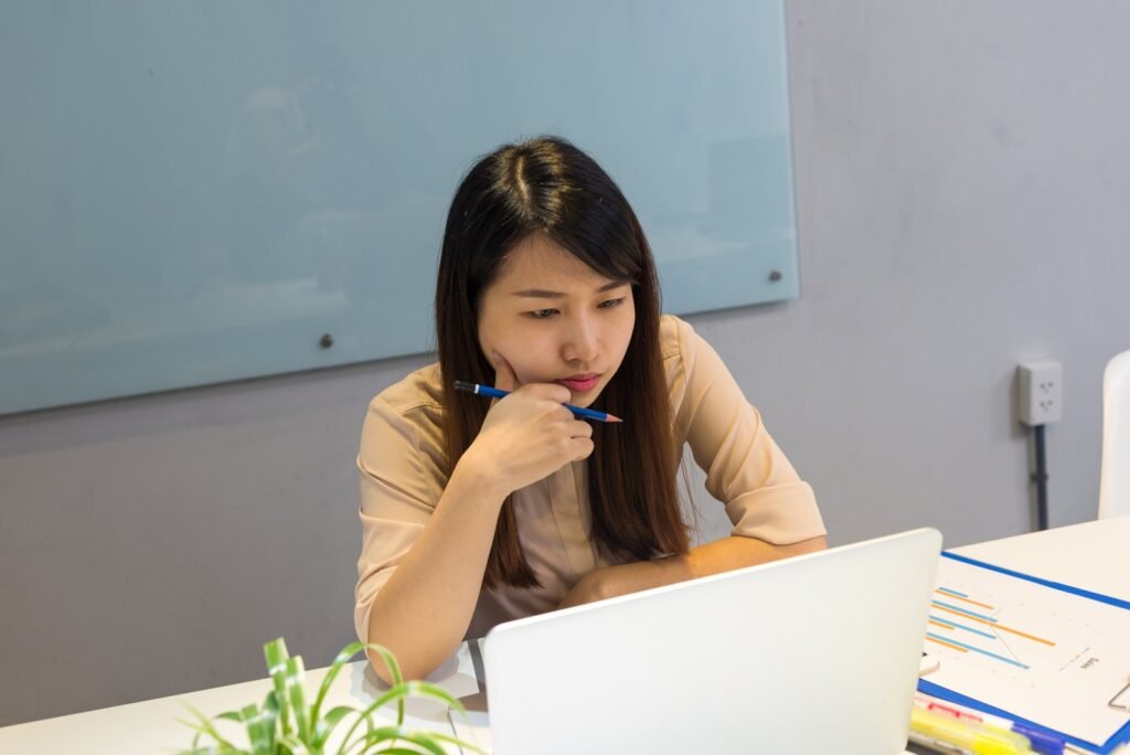 Office worker holding a pen to her chin while carefully reading a laptop screen