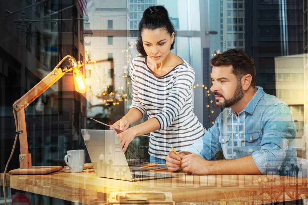 Two colleagues at a desk, one showing the other a task on a laptop